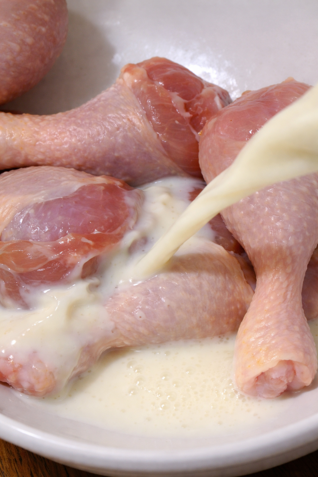 Raw chicken drumsticks are being marinated in fresh milk in a bowl.
