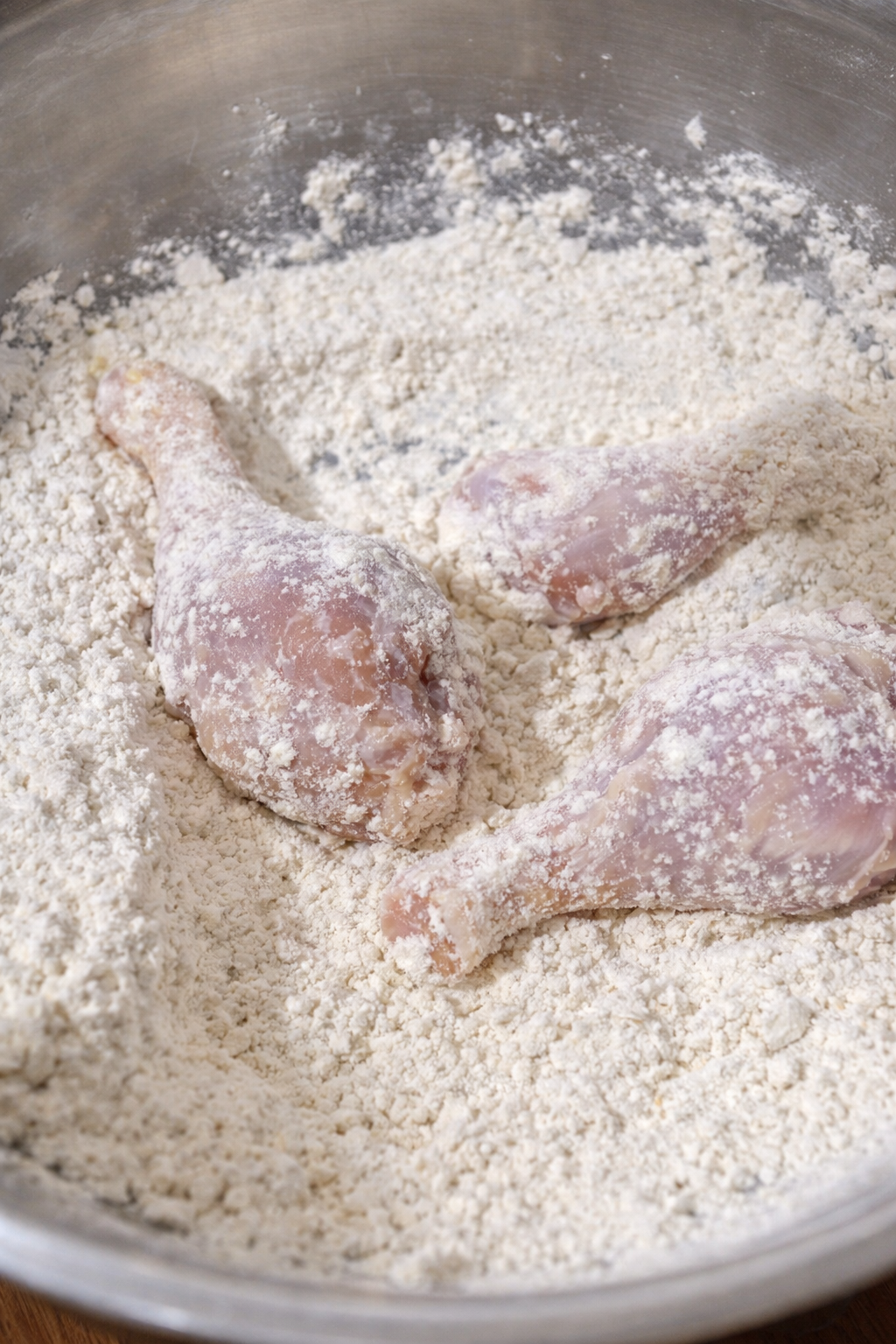 Raw chicken drumsticks are being coated in seasoned flour for frying.