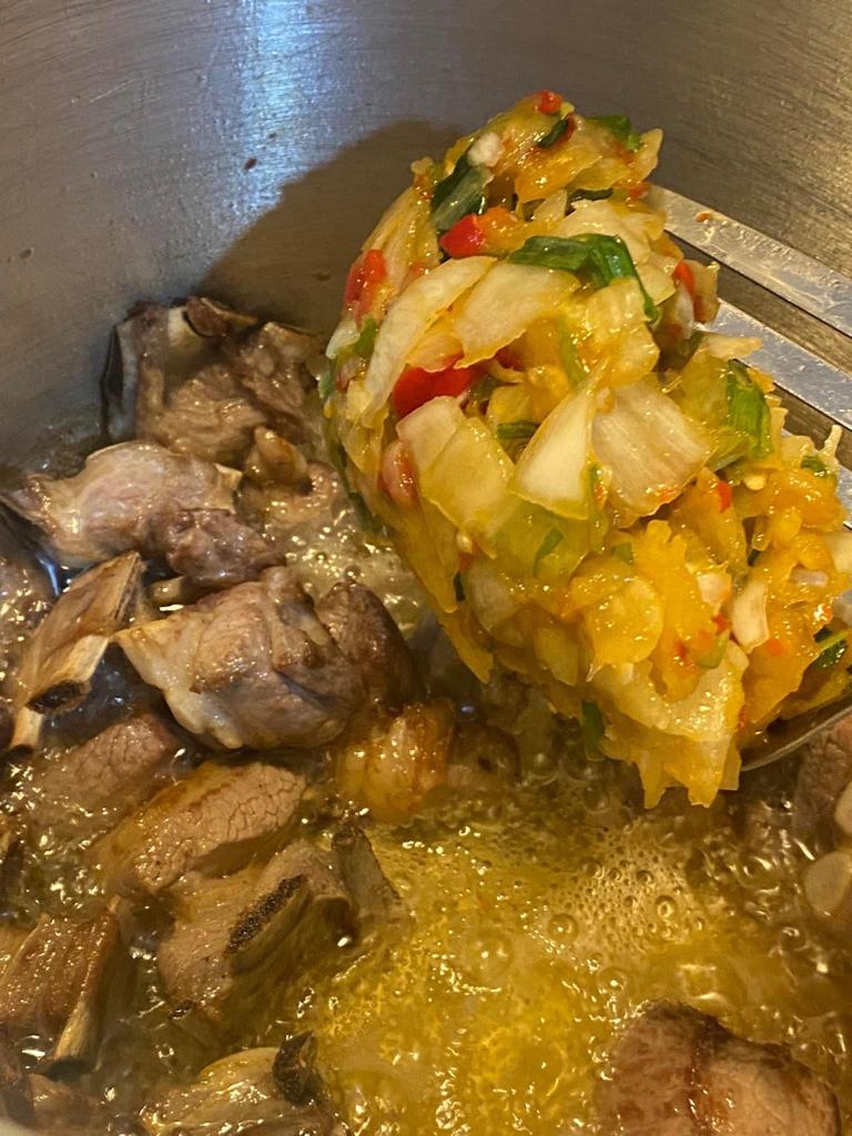 Spoonful of blended onions, spring onions, and peppers being added to beef stew in a pot.