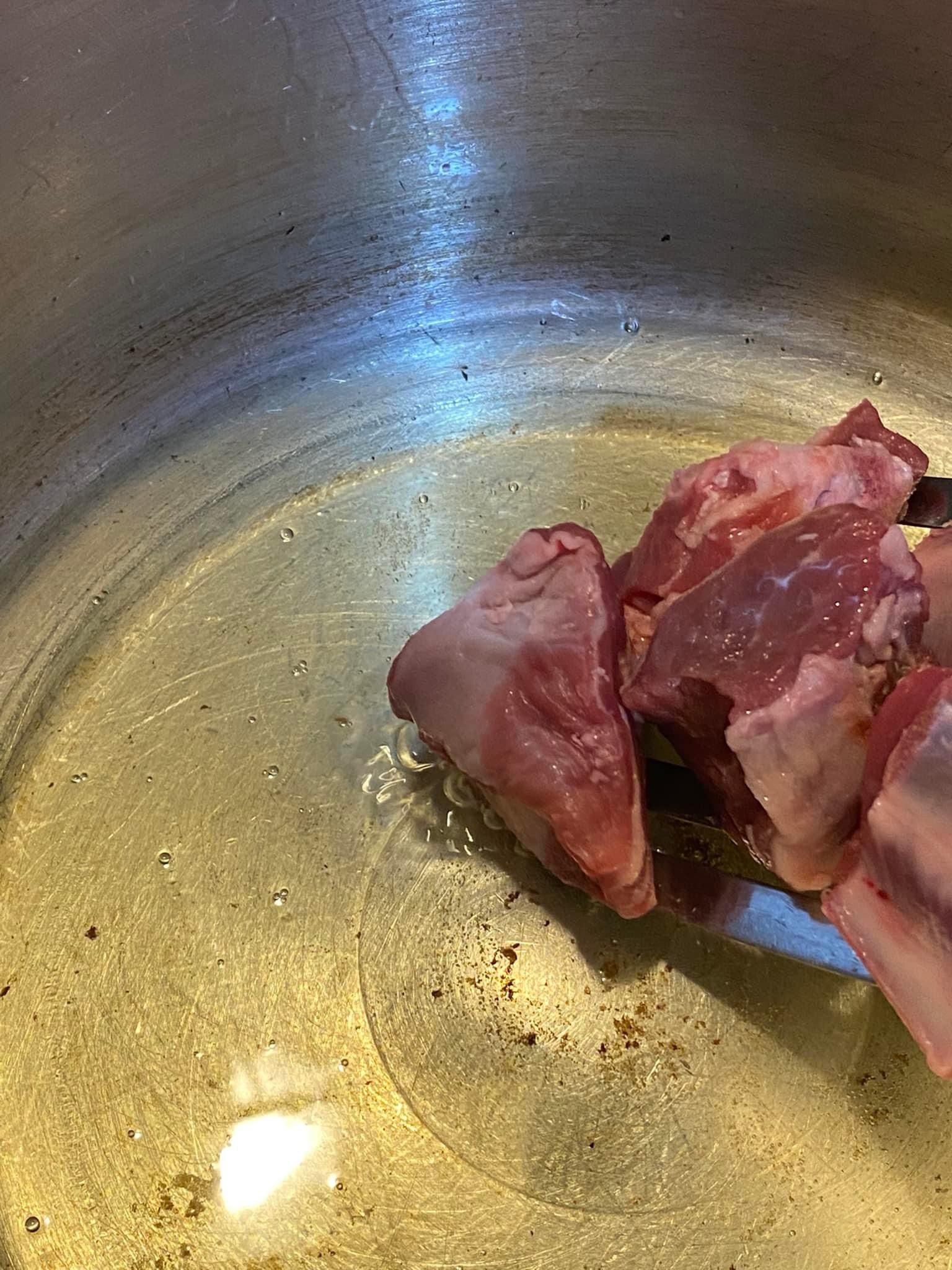 Raw chunks of lamb and beef beginning to brown in hot oil inside a stainless steel pot.