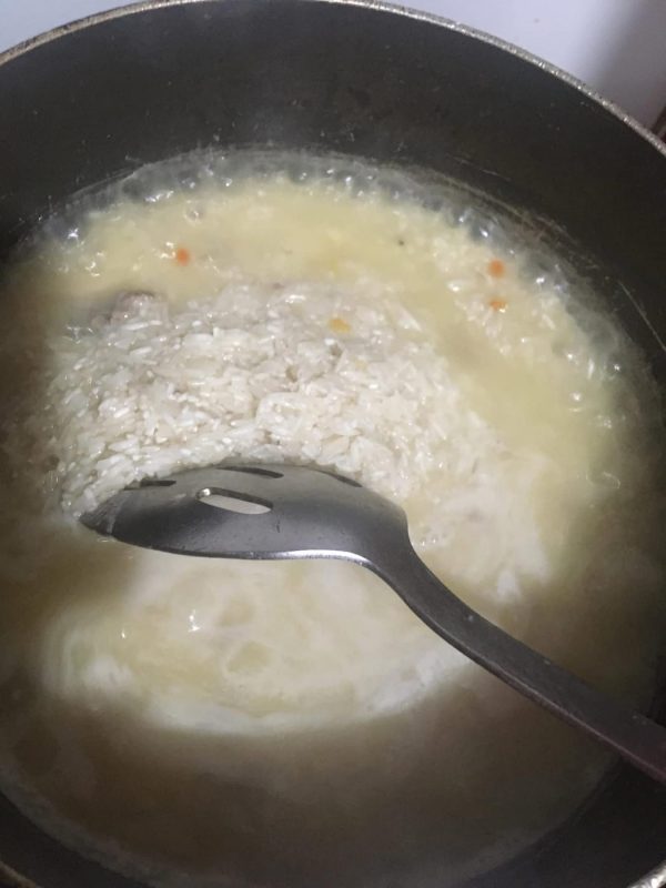 A close-up view of rice simmering in a yellow, flavorful chicken broth inside a pot. A metal spoon rests in the pot, partially submerged in the softening rice, while bubbles rise from the gently boiling broth.