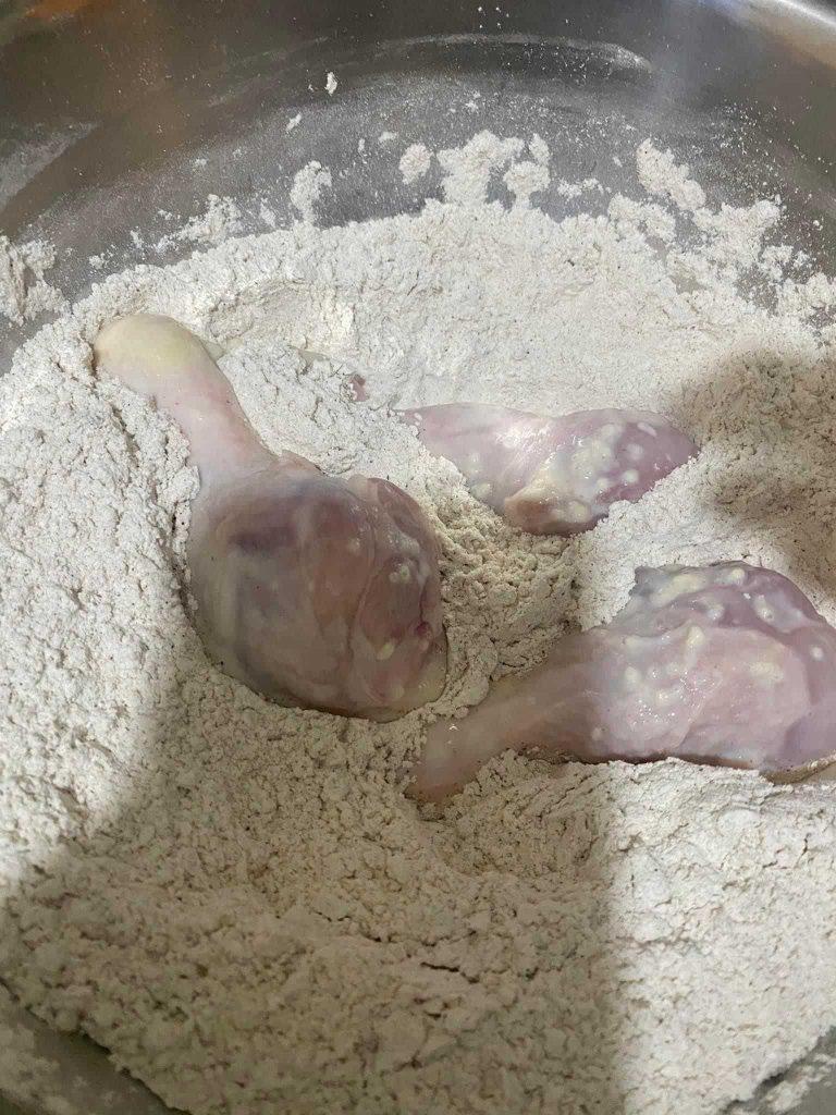 Raw chicken drumsticks are being coated in seasoned flour for frying.