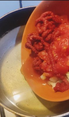 Tomato paste and chopped tomatoes are being added to onions in a saucepan with oil