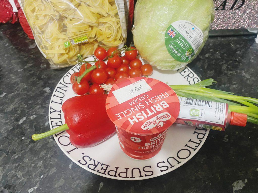 A colorful arrangement of ingredients for creamy chicken pasta, including pasta, lettuce, cherry tomatoes, red bell pepper, cream, spring onion, and tomato paste, all placed on a decorative plate.