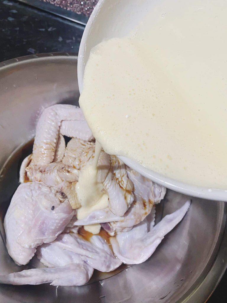 Pouring cornflour batter over seasoned raw chicken wings in a metal bowl.