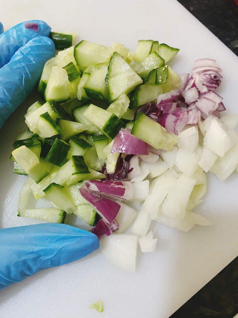 Chopped cucumber, red onion, and white onion on a white chopping board with a gloved hand