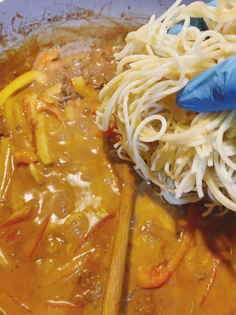 Drained spaghetti being added to a creamy beef and pepper sauce in a pot, with a gloved hand holding the pasta.