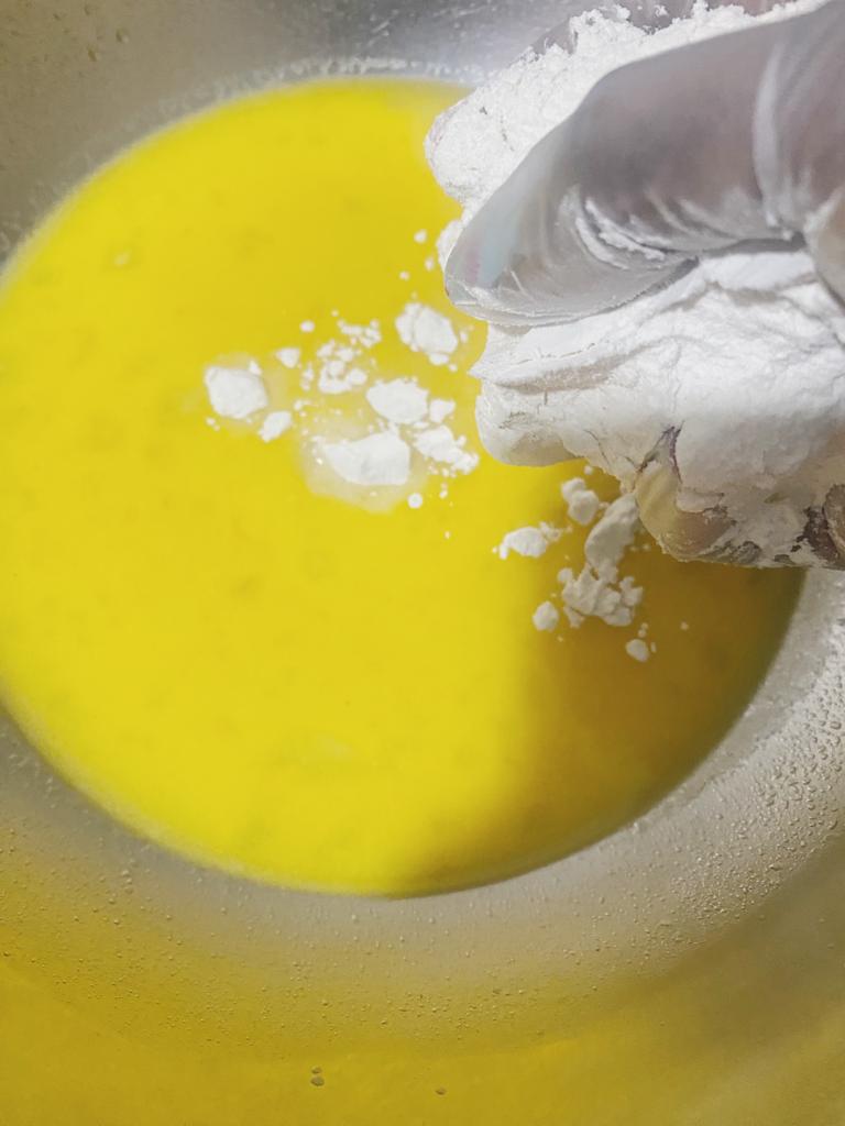 Flour is being added by hand into a bowl of yellow dough mixture.