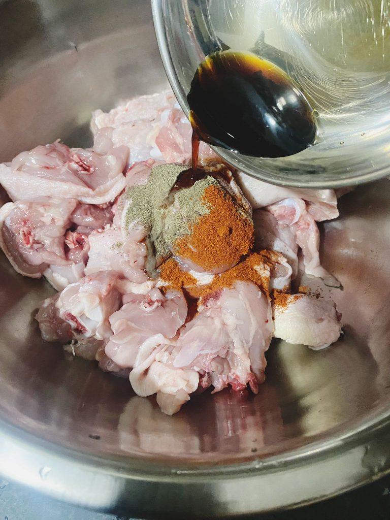 Marinade being poured over raw chicken pieces in a large silver bowl, featuring spices like paprika and garlic paste.