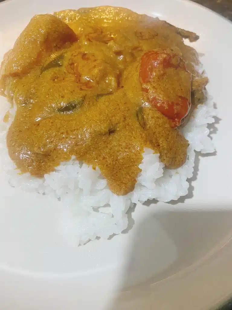 A plate of steamed white rice topped with a creamy, golden-orange stew featuring tender pieces of meat, whole tomatoes, and rich spices.