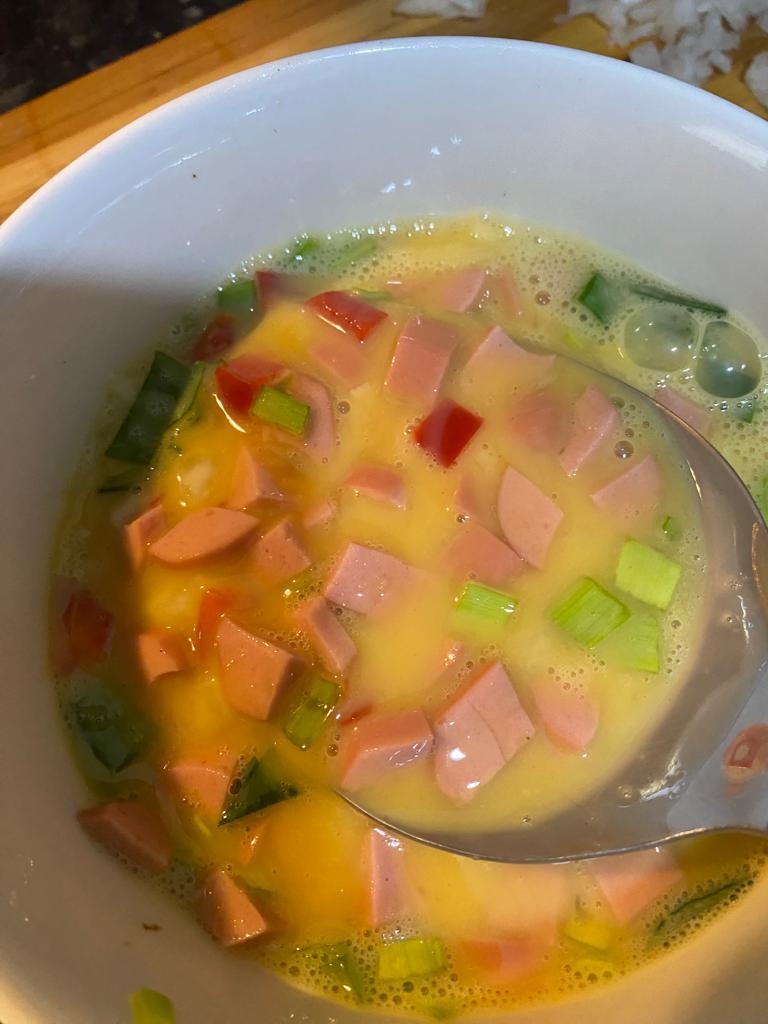 A mixing bowl filled with beaten eggs combined with chopped sausages, red and green paprika, and onions. The colorful ingredients float in the egg mixture, creating a vibrant base for a savory omelet.