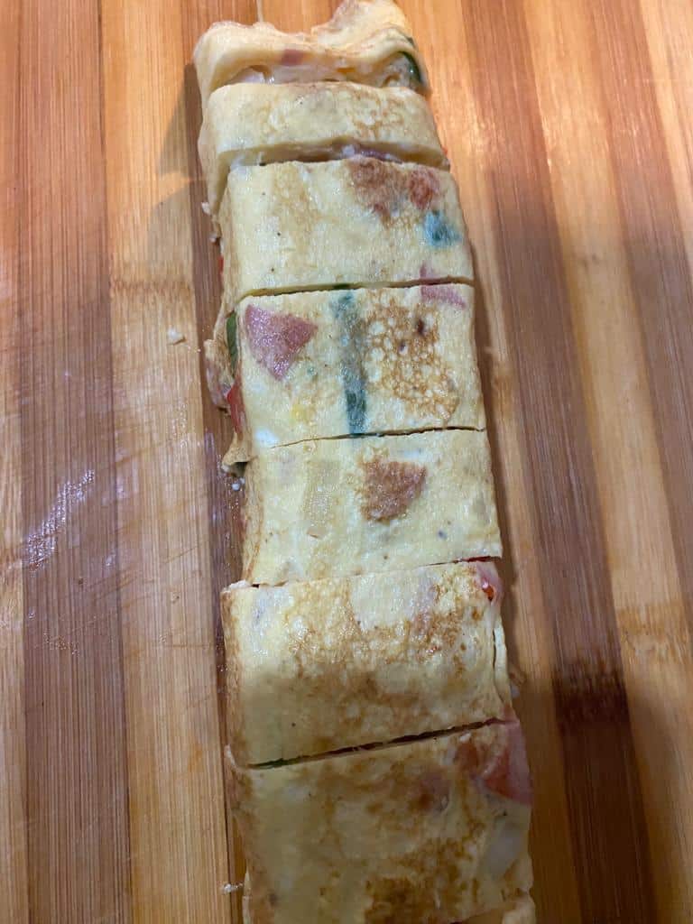 Rolled omelet sliced into pieces on a cutting board, showing golden egg layers filled with sausage and red and green paprika