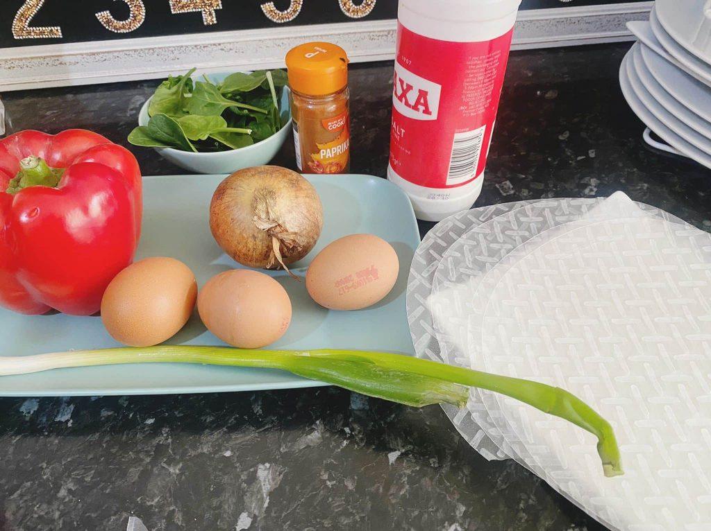 Ingredients including beaten eggs, chopped onion, paprika powder, fresh spinach, rice paper sheets, chopped spring onion, and oil ready for frying.