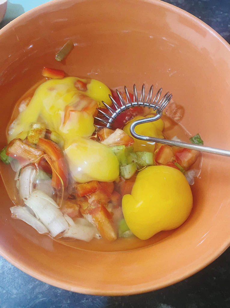 A mixing bowl containing beaten eggs with diced onions, spring onions, salt, and paprika being combined to prepare an omelette.