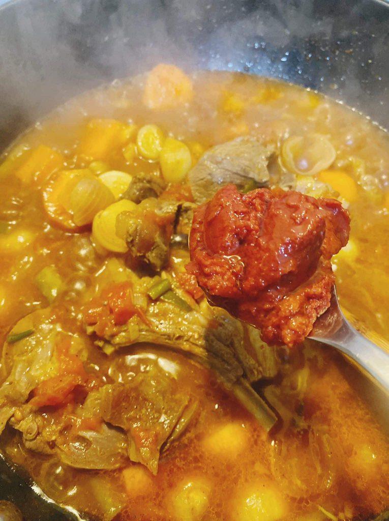 A rich soup simmering with chunks of vegetables and a spoonful of tomato paste ready to be stirred in.