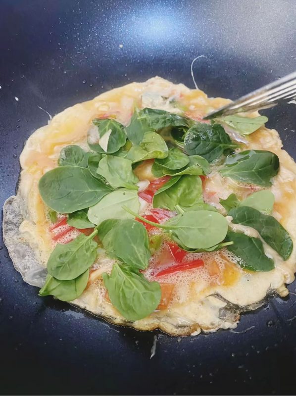 Egg mixture cooking on rice paper with fresh spinach added on top in frying pan