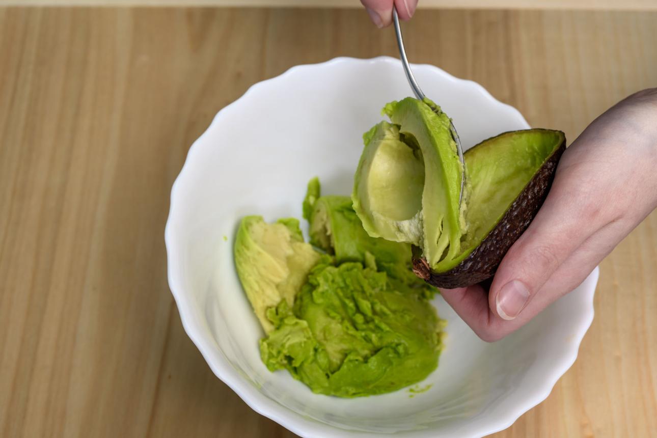 Scooping avocado flesh into a small bowl with a spoon