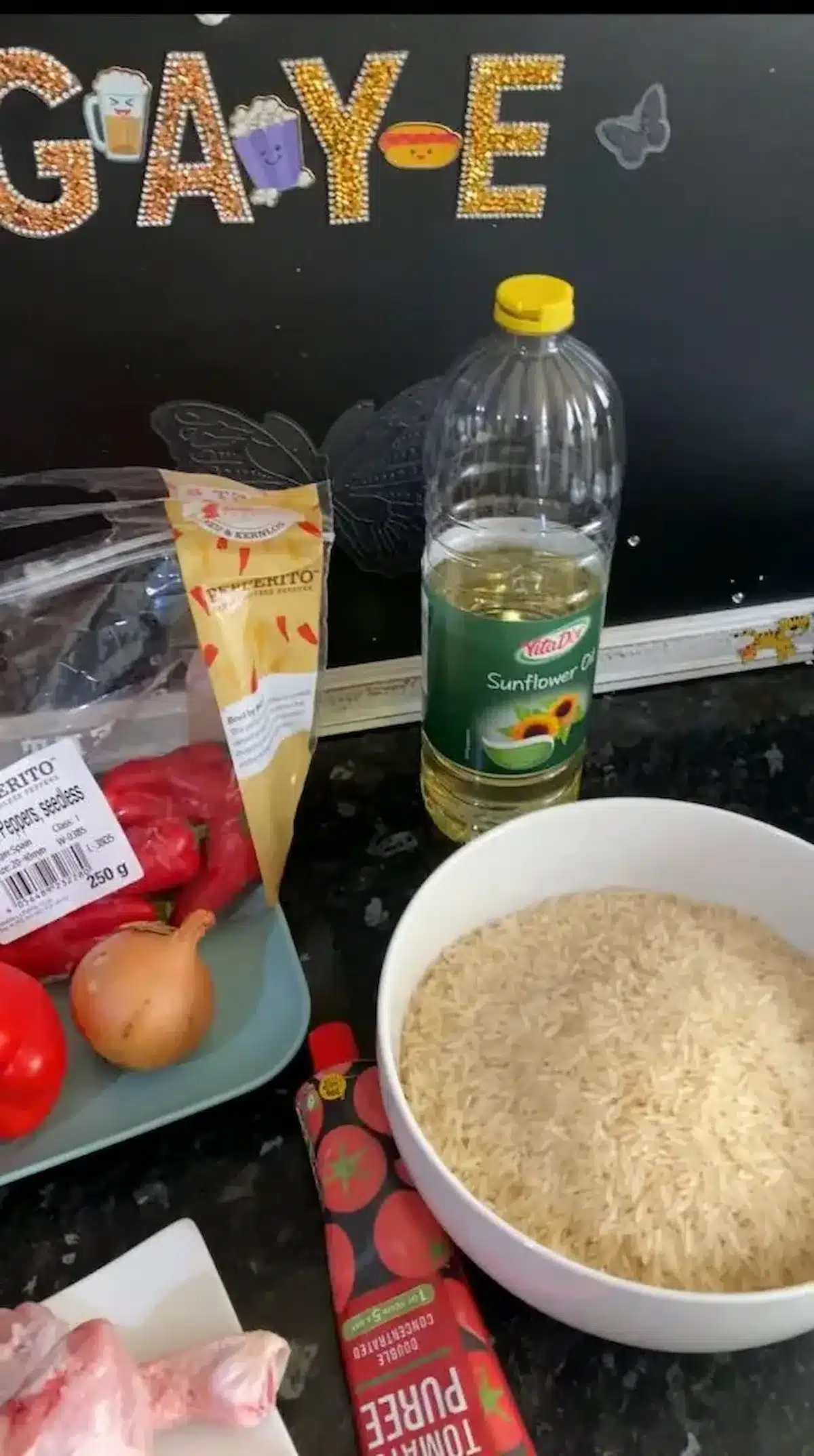 A cooking preparation setup featuring sunflower oil, uncooked rice in a white bowl, tomato puree, an onion, red peppers, and chicken drumsticks, all arranged on a black countertop with a decorative 'GAYE' sign in the background.