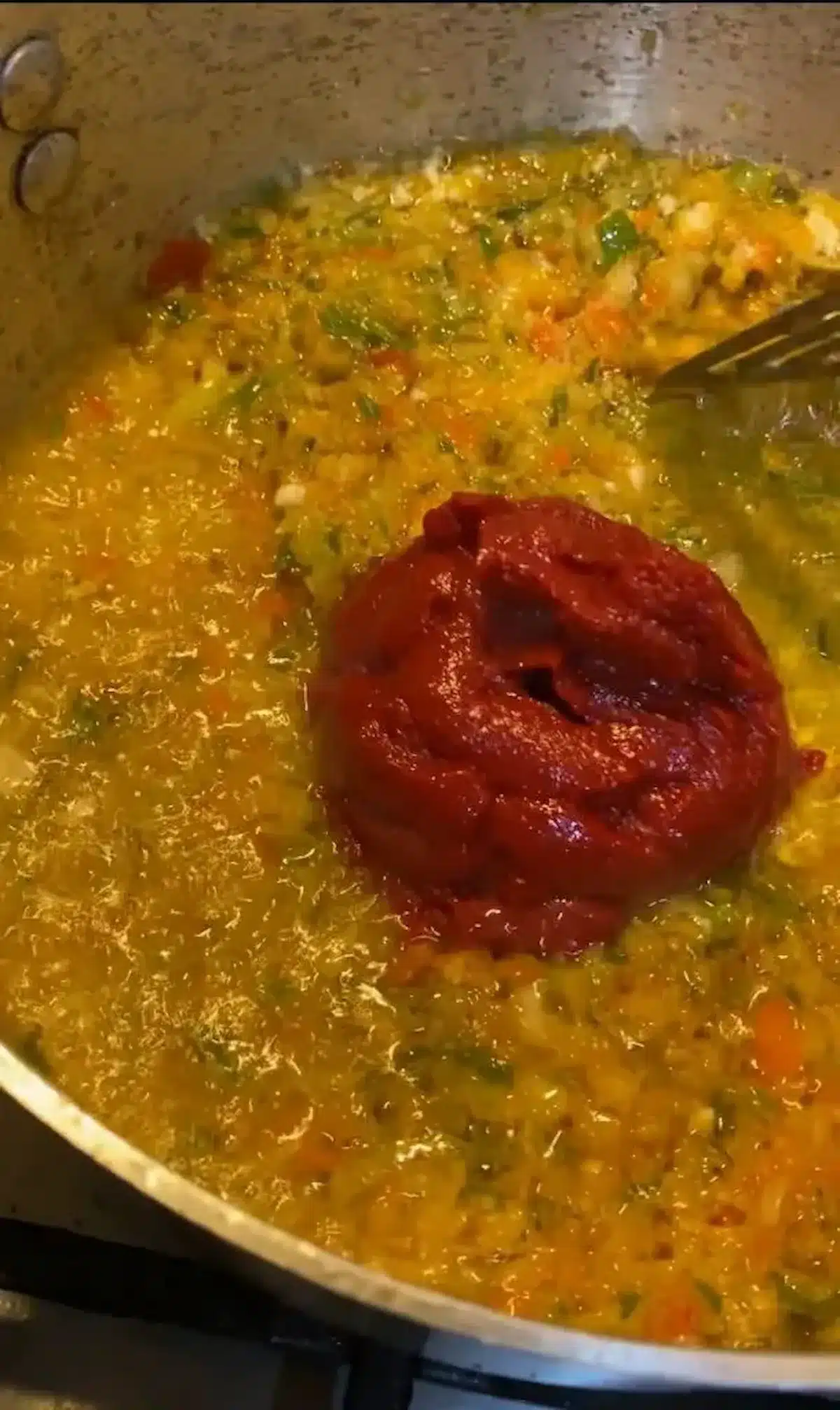 A pan filled with a vibrant mix of yellow vegetable sauce, chopped green peppers, and a dollop of rich red tomato paste being stirred during the cooking process.