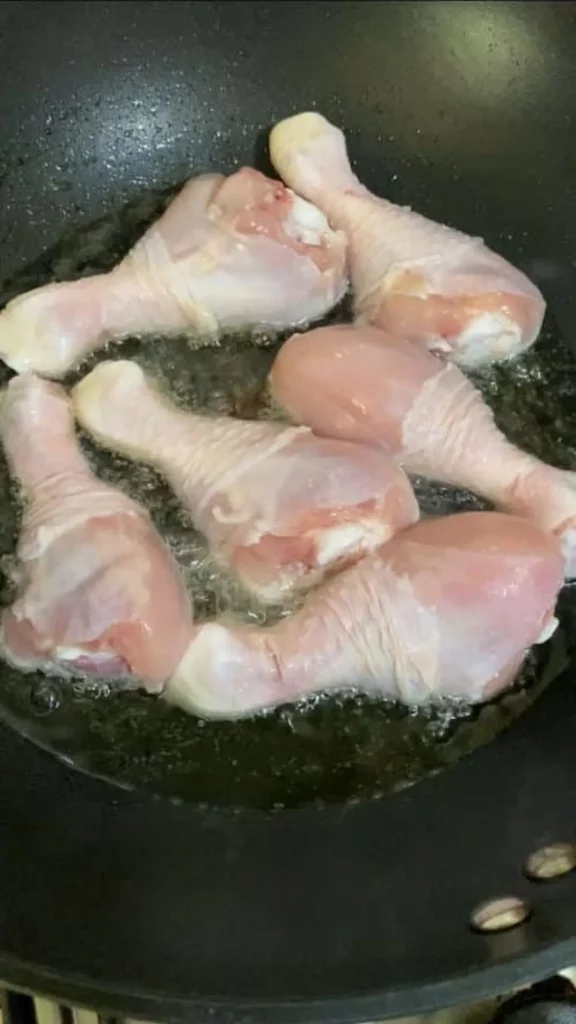 Six raw chicken drumsticks frying in a hot pan of oil, beginning to turn golden brown, as part of the cooking process.