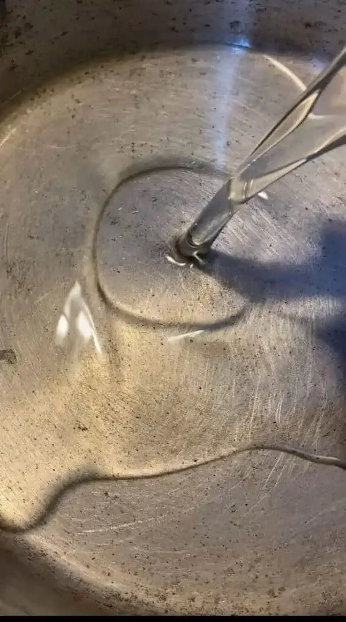 A close-up of oil being poured into a large, well-used silver cooking pot, showing a reflective surface with natural wear marks.