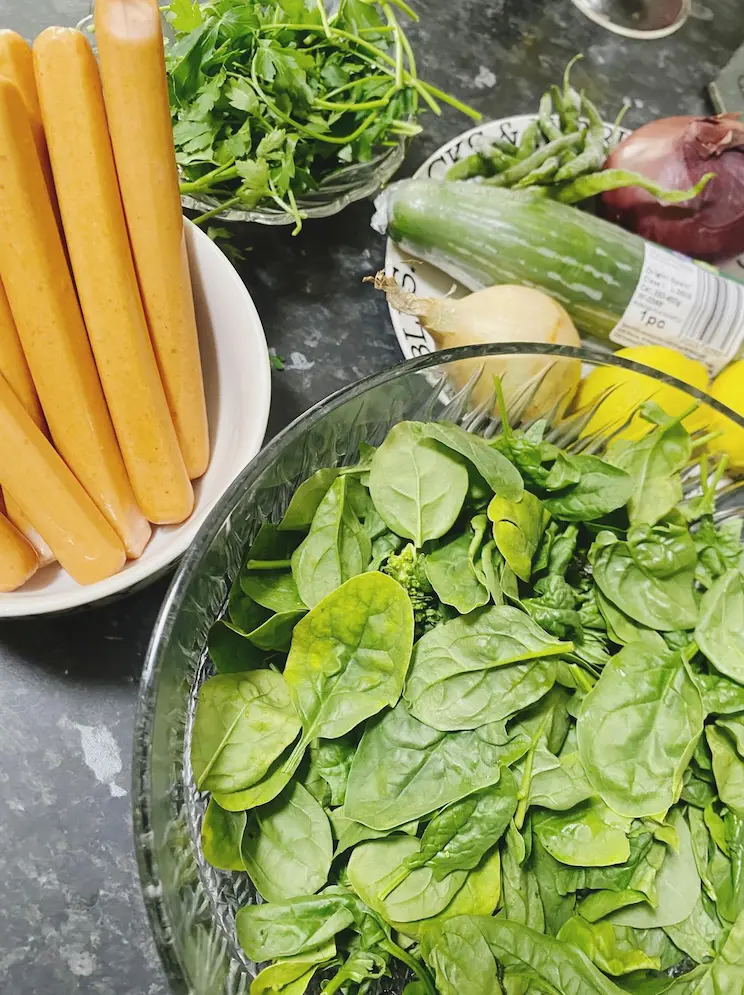 Fresh spinach, sausages, parsley, cucumber, onions, green chilies, and lemons arranged on a kitchen counter before cooking.