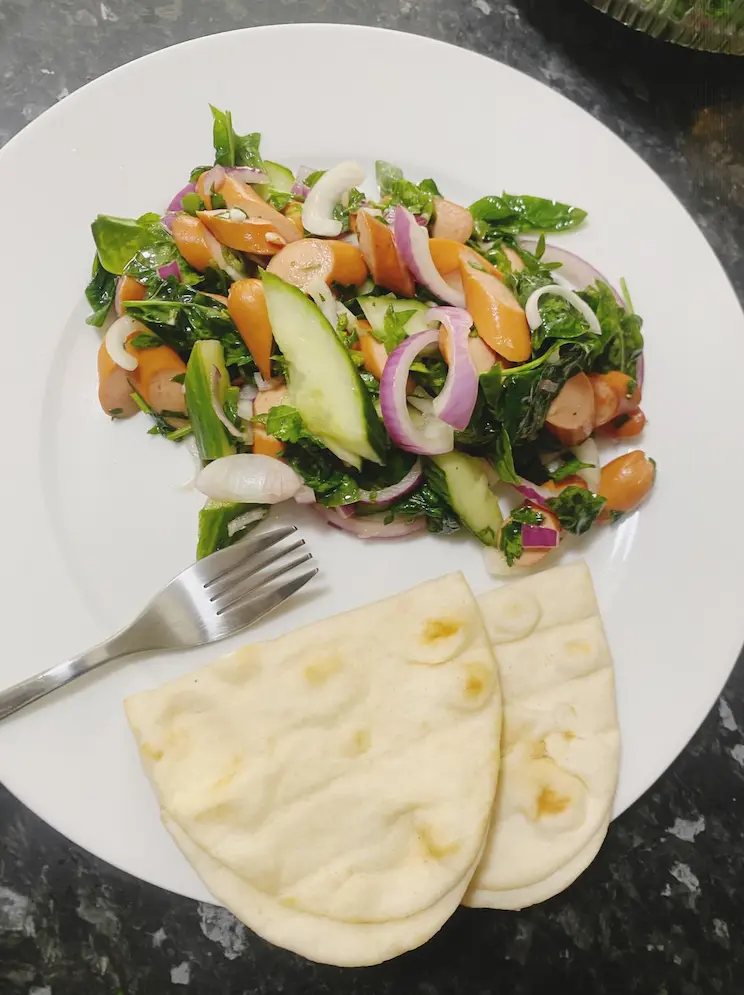 Fresh spinach and sausage salad served on a white plate with warm naan bread.