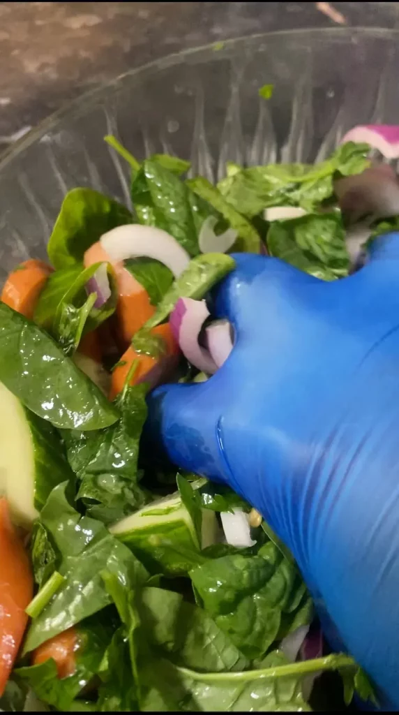 A hand wearing a blue glove mixing spinach, sausage, onion, cucumber, and chili in a large bowl.