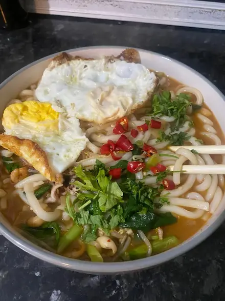 A homemade noodle bowl topped with fried eggs, fresh herbs, and chilies, representing Tima’s comforting, multicultural cooking style.